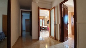 Corredor que dá para uma sala de jantar com mesa e cadeiras, uma porta à direita e, no quarto à esquerda, uma cama com cobertura estampada.