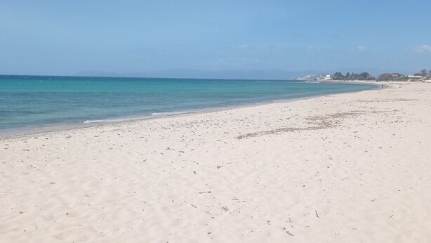 Uma extensa praia de areia clara com águas azul-turquesa, alguns edifícios distantes e céu limpo.