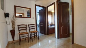 Um corredor com duas cadeiras de madeira, uma planta em vaso, um espelho e uma porta que dá para um quarto.