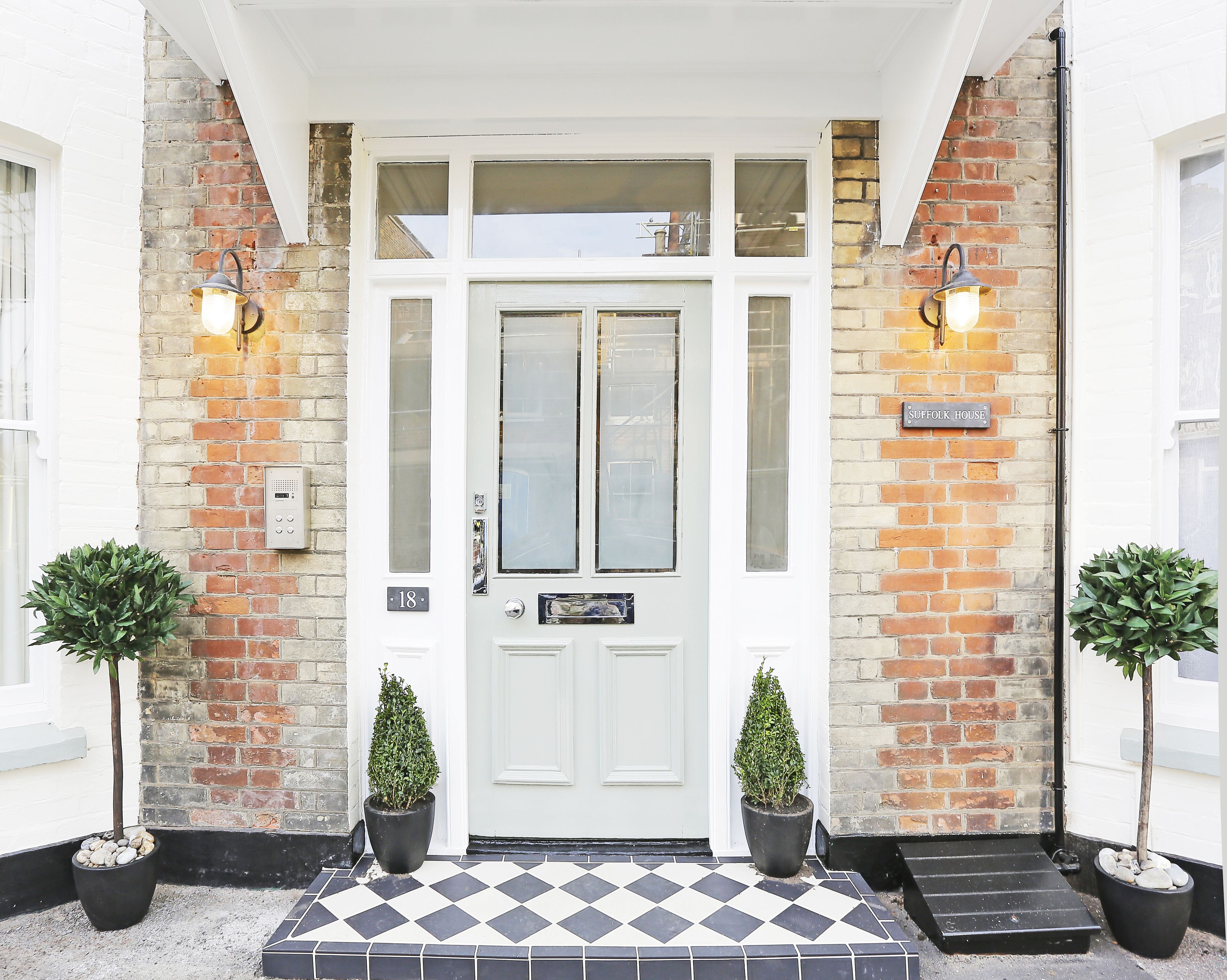 A white double door with a glass panel, a brick wall, and a black and white checkered entrance. There are two potted plants on either side of the door.
