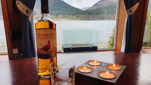 A bottle of Famous Grouse whisky, a glass, and four lit candles on a table with a scenic lake view. - Waterfront with Private Hot Tub, Kayaks and Paddleboards free for guests use !!! (ARROCHAR)