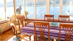 A dining area with a table and chairs, large windows with a view of a lake and mountains, and a yellow umbrella. - Waterfront with Private Hot Tub, Kayaks and Paddleboards free for guests use !!! (ARROCHAR)