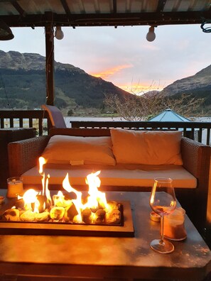 Un área de estar al aire libre acogedora, con una chimenea y una copa de vino, con vistas a un paisaje montañoso.