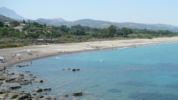 Une plage à l’eau d’un bleu limpide, avec un rivage rocheux et des montagnes qui se dessinent à l’horizon.