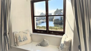 A room with a window overlooking a harbor, a white bench with cushions, and curtains.