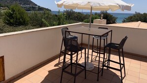 Una terraza con mesa y sillas, un paraguas y vistas al mar y a las colinas.