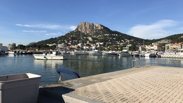 Una marina con barcos amarrados, una colina rocosa al fondo y un paseo pavimentado en primer plano.