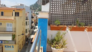 Una calle estrecha flanqueada por edificios, una fuente de agua azul y macetas con plantas en los balcones.