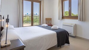 A hotel room with a bed, a chair, a table with a lamp, and a radiator. There are two windows with curtains, and a balcony is visible through one window. - Ca Na Volantina - Great family villa close to Puerto Pollensa (Port de Pollença)