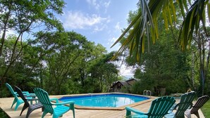 Une terrasse en bois avec une piscine, entourée de chaises vertes et d’une végétation luxuriante.