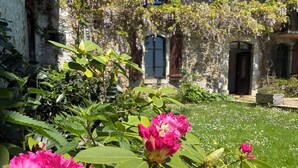 Un bâtiment en pierre avec un jardin devant, où s’épanouissent des fleurs roses et une végétation verdoyante.