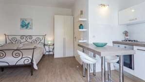 A compact bedroom with a bed, bedside table, and a small desk with a chair. There is a kitchenette with a stove and a sink. - Studio Apartment Klara  Breathtaking view to the sea and Old Town! (Dubrovnik)