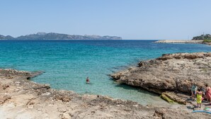 Une côte rocheuse bordée d’une eau turquoise et limpide, une personne qui nage et un groupe de personnes sur la plage.