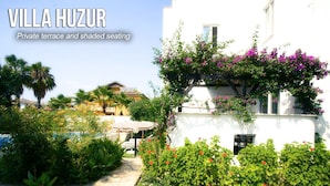 A villa with a private terrace and shaded seating area, surrounded by lush greenery and flowering plants.