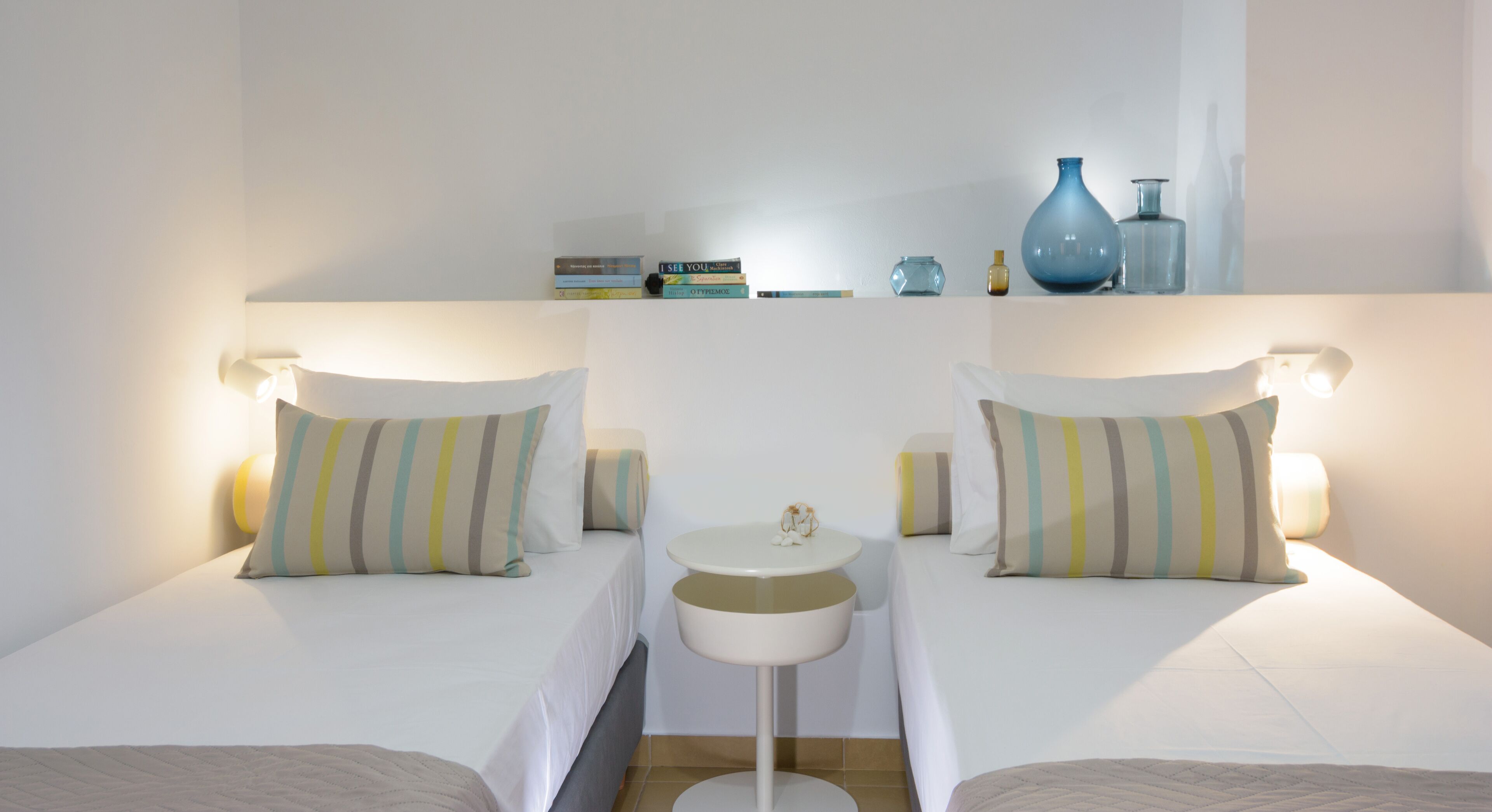 Een hotelkamer met twee bedden, een klein rond tafeltje en een plank met boeken en decoratieve objecten.