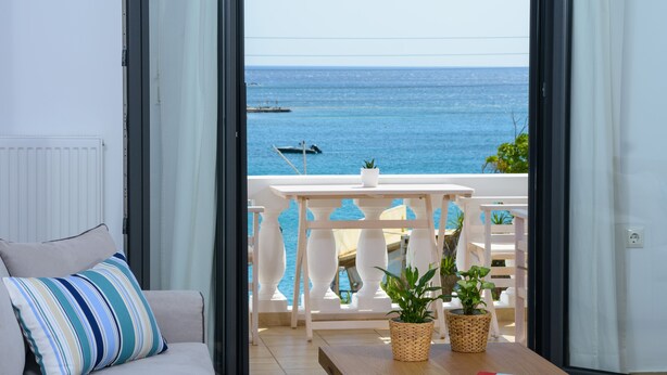 A balcony with a view of the sea, a white sofa with striped pillows, and two potted plants on a wooden table. - Summer Breeze! Airy, spacious and fully equipped sea view apartment. (Ag. Nikolaos)