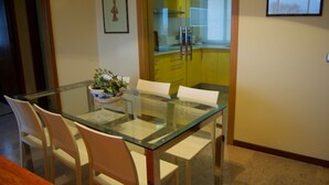 Un comedor con una mesa de vidrio y sillas blancas, un cuenco de fruta y un armario amarillo al fondo.