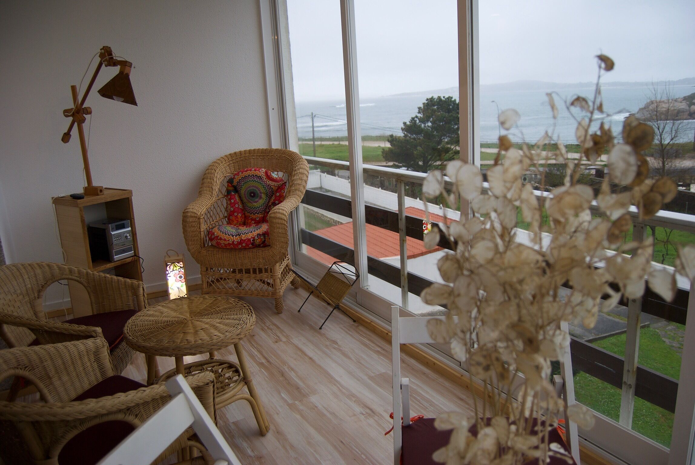 Une pièce meublée de mobilier en osier, une petite table avec une lampe et une vue sur l’océan par de grandes fenêtres.