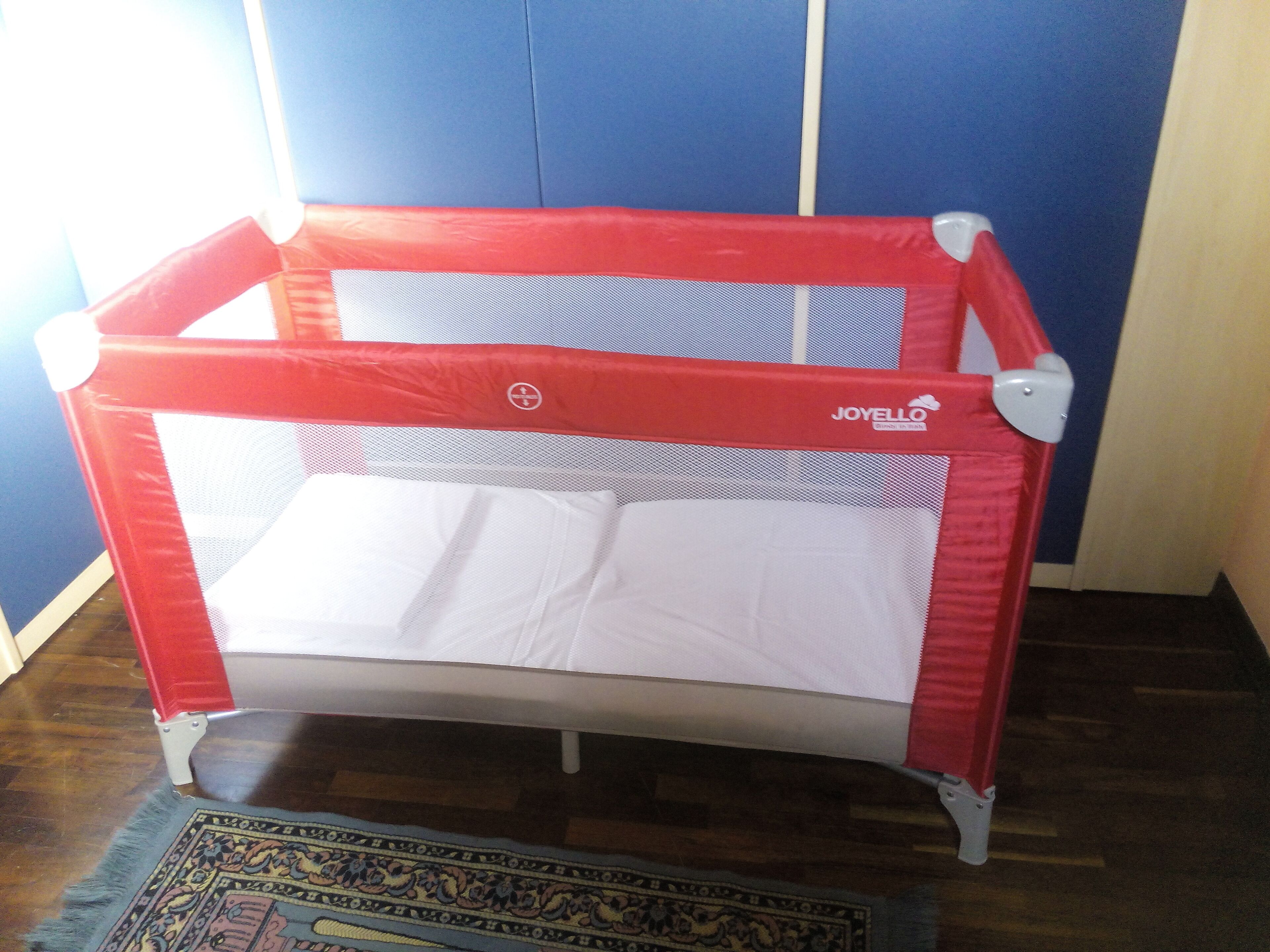 Un parc pour bébé rouge, doté d’une partie supérieure en tissu à mailles et d’un matelas blanc, est placé sur un plancher en bois, sur lequel repose un tapis à motifs.