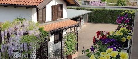 Una zona residencial con tejados de tejas de arcilla, un edificio blanco con contraventanas de madera y un jardín con flores púrpuras y amarillas.