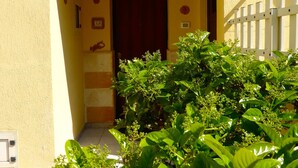 A yellow building with a red door, a white fence, and green plants in front.