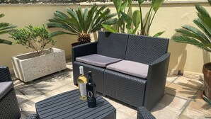 A wicker sofa with cushions, a small table with a bottle of wine, and potted plants on a tiled patio.