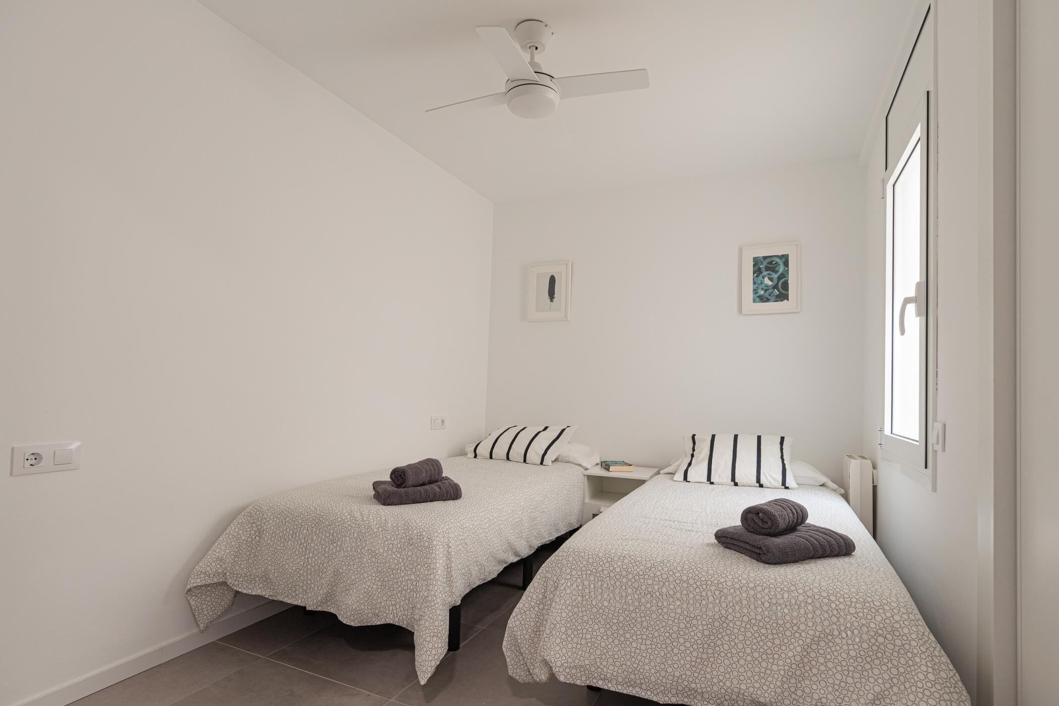 Ein Zimmer mit zwei Betten, einem Deckenventilator, einem kleinen Fenster und einem gerahmten Bild an der Wand.