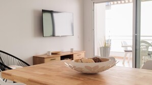 A modern dining area with a wooden table, white chairs, a white bowl with pastries, a flat-screen TV on the wall, and a sliding glass door leading to an outdoor area.