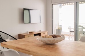 A modern dining area with a wooden table, white chairs, a white bowl with pastries, a flat-screen TV on the wall, and a sliding glass door leading to an outdoor area. - WHITE APARTMENT with TERRACE, AC & PARKING, NEAR BEACH (Tossa de Mar)