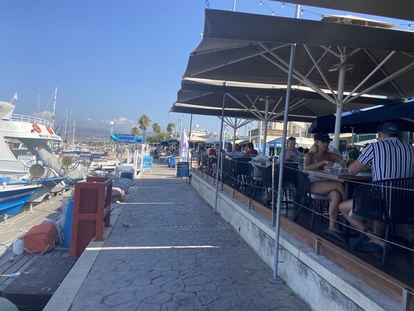 A marina with boats docked, outdoor seating under umbrellas, and a sign for Blue Lagoon tickets. - 3 Bedroom Villa With Private Pool & Sea Views (Kamares Village)