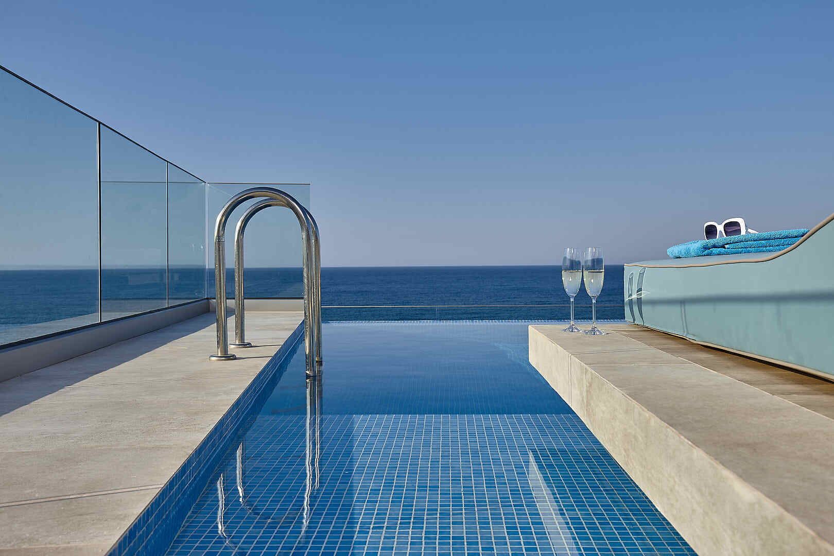 Ein Infinity-Pool auf dem Dach mit freiem Blick auf den Ozean, einer Glastrennwand und zwei Champagnergläsern auf einer gepolsterten Sitzgruppe.