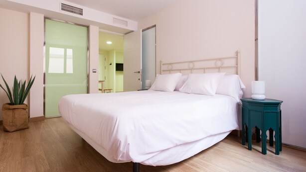 Een slaapkamer met een bed, nachtkastje, een plant in pot en een deur naar een andere kamer.