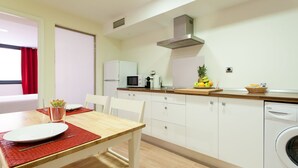 Een kleine kitchenette met een magnetron, broodrooster en wasbak. Er staat een eettafel met stoelen en een rode placemat. Een wasmachine is in de hoek te zien.