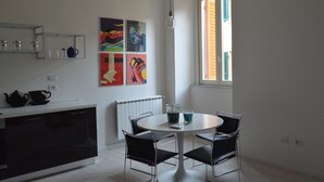 A modern kitchen with a dining area, featuring a white countertop, black cabinets, and a round table with four chairs. There are colorful paintings on the wall, a window with shutters, and a hanging light bulb. - Apartment in the historic center of Perugia (Perugia)