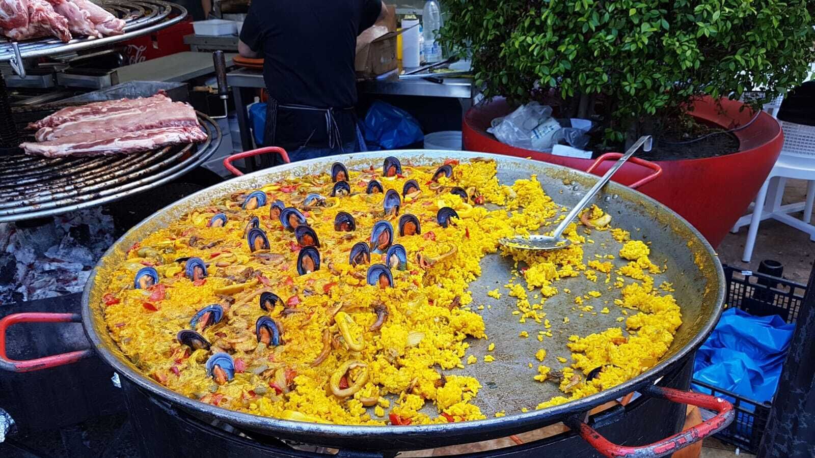 Una grande paellera piena di riso giallo e frutti di mare, posizionata all'aperto con una griglia e piante in vaso sullo sfondo.