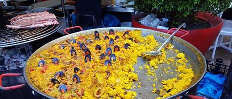 Una grande paellera piena di riso giallo e frutti di mare, posizionata all'aperto con una griglia e piante in vaso sullo sfondo.
