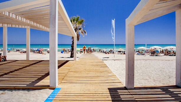 A wooden boardwalk leading to a beach with white structures and palm trees. - Apart. RIVERA II, location in Old town and only 10 minutes the beach. (Alicante)