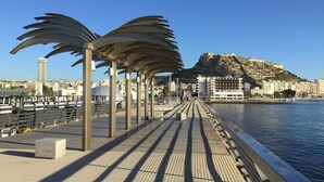 A waterfront promenade with palm-like structures, a clear view of the sea, and a distant hill with buildings. - Apart. RIVERA II, location in Old town and only 10 minutes the beach. (Alicante)