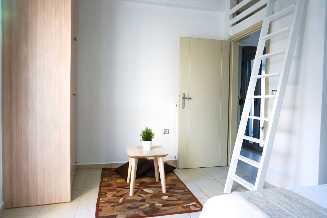 A room with a wooden wardrobe, a small table with a potted plant, a white door, and a white ladder leaning against the wall.