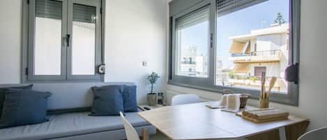 A modern living room with a sofa, a dining table, and a view of buildings through large windows.
