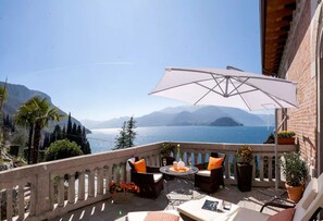 A balcony with wicker furniture, a table with a white umbrella, and a view of the sea and mountains.