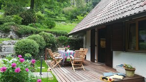 Una terraza de madera con una mesa y sillas, rodeada de abundante vegetación y un muro de piedra.