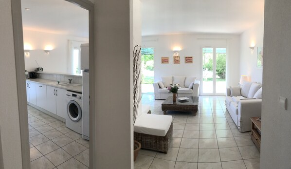 A modern, well-lit living area with a sofa and a coffee table, a kitchen with a washing machine, and a view of greenery outside.