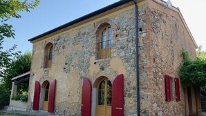 A stone building with two arched windows and red shutters, featuring a yellow door and a small balcony.