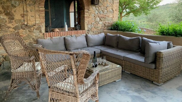 A stone-walled patio with a wicker sofa and chairs, a small table with wine glasses, and a view of greenery.