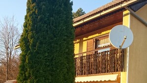 Un edificio de dos pisos con un balcón, una antena parabólica y un alto pino al frente.