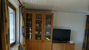 Una habitación con armarios de madera, televisor, un reposapiés amarillo y una ventana con cortinas.