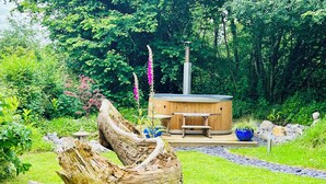 A hot tub surrounded by a wooden deck, with a rustic tree stump in the foreground and lush greenery in the background.