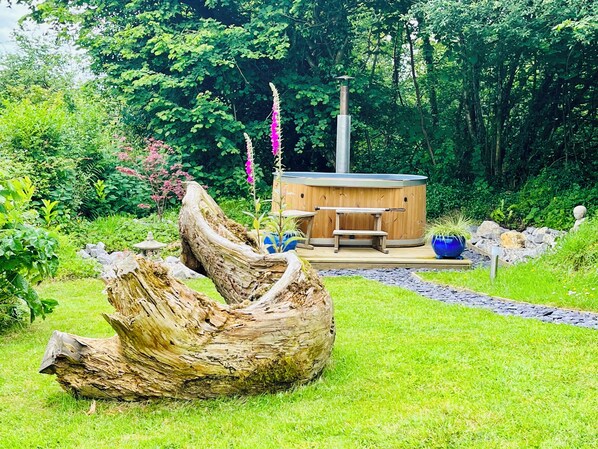 A hot tub surrounded by a wooden deck, with a rustic tree stump in the foreground and lush greenery in the background.