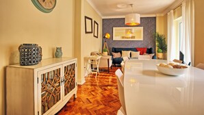 Una sala de estar moderna con comedor, que cuenta con piso de madera en diseño espiga, una mesa de comedor blanca y un sofá con un diseño decorativo en la pared de fondo.